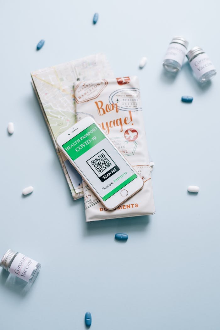 A high-angle shot of a smartphone displaying a health passport with COVID-19 QR code, surrounded by vaccine vials, pills, and travel documents.